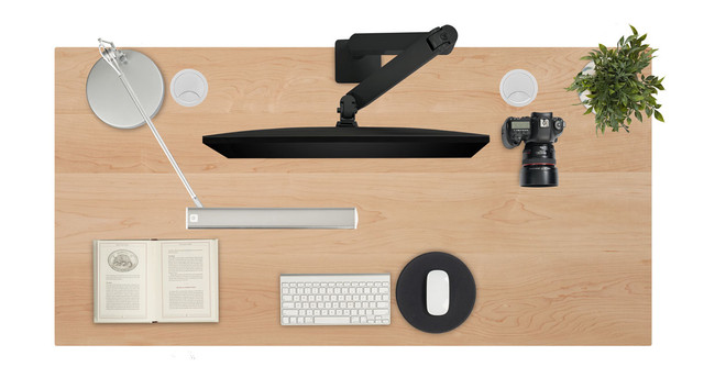 A tidy desk setup with a vertical monitor, keyboard, mouse, camera, notebook, lamp, and plant on a wooden desk.