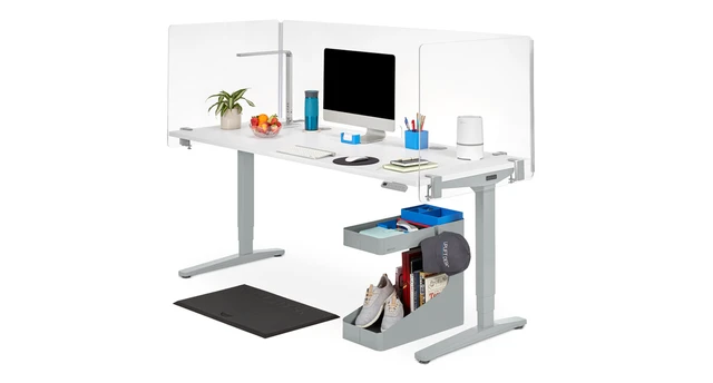 Another white office desk with a built-in cable organizer, transparent side shields, keyboard, mouse, and storage bin with books and shoes underneath