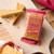 Golden, buttery oat flapjack bar wrapped in Lottie Shaw’s signature pink and gold packaging, resting on a wooden board with crumbly flapjack wedges scattered around