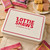 Cream and red Lottie Shaw’s tin on rustic wooden table with flapjack bars and millionaires shortbread