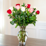 One dozen premium red, pink, and white long-stemmed roses arranged in a classic glass urn style vase with bright greenery photographed on a dark wooden table with light shinning in through a window in the back. 
