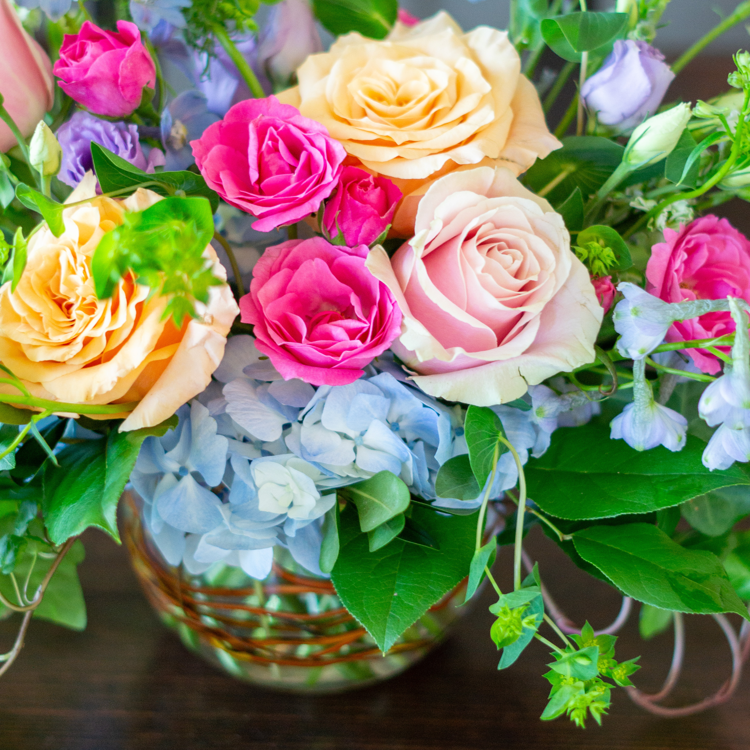 Peach valentines day flowers on a blue tablecloth. Flowers include peach roses, cream lisianthus, peach stock, white hydrangea, pink spray roses, and pink hypericum berries accented with eucalyptus. 