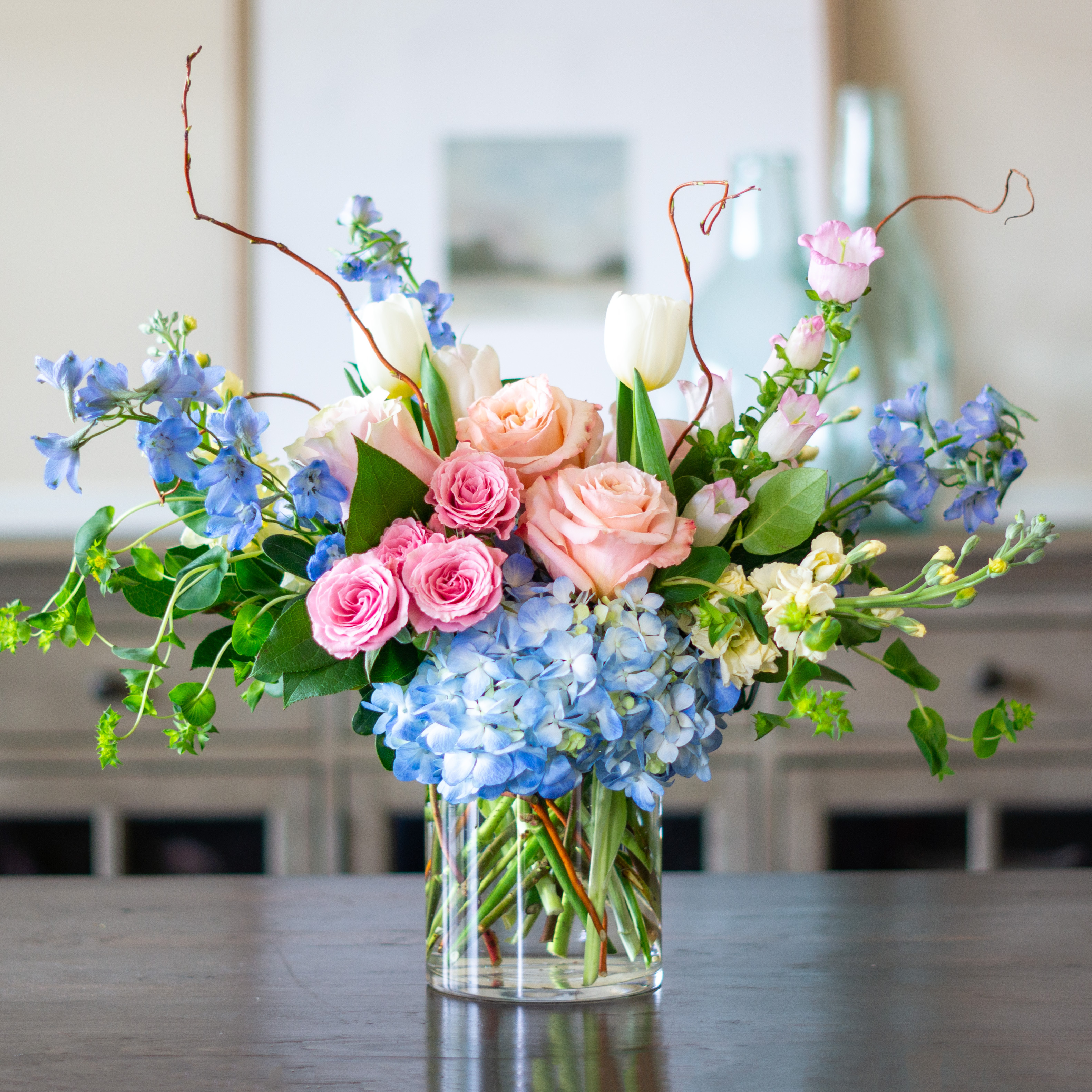 Peach valentines day flowers on a blue tablecloth. Flowers include peach roses, cream lisianthus, peach stock, white hydrangea, pink spray roses, and pink hypericum berries accented with eucalyptus. 