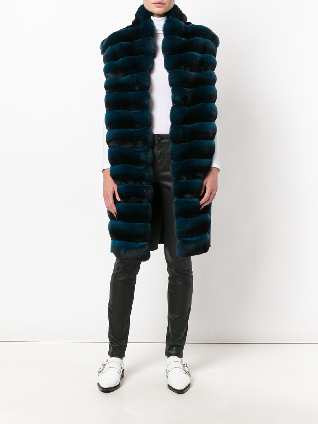 long green chinchilla fur vest  ,with stand up collar on model wearing white turtleneck blouuse, black leather pants and white leather shoes with straps