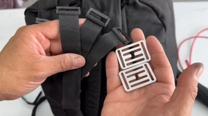 Hand holding two metal H-shaped tabs next to a black backpack strap and velcro straps.