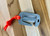 Compact stage rigging safety cutter with an orange cord lanyard on a wooden surface, showing hook-style design and recessed blade.