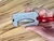 Side view of a metal utility blade diver’s cutter held in hand with a red paracord lanyard attached, showing the elongated cutting slot.