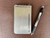 Small spiral notebook with a brushed titanium cover engraved with a name, placed next to a pen on a textured surface.