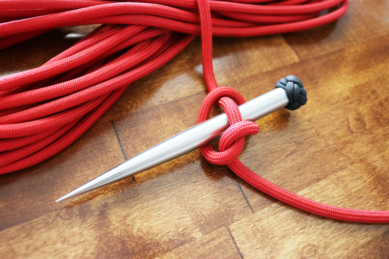 Stainless steel marlinspike tool used with red paracord tied around the spike for knot tying and rope work on a wooden surface.