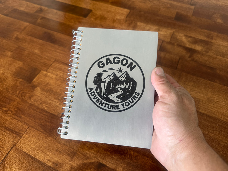 Brushed aluminum notebook cover with a custom engraved logo on the front, held in a hand.