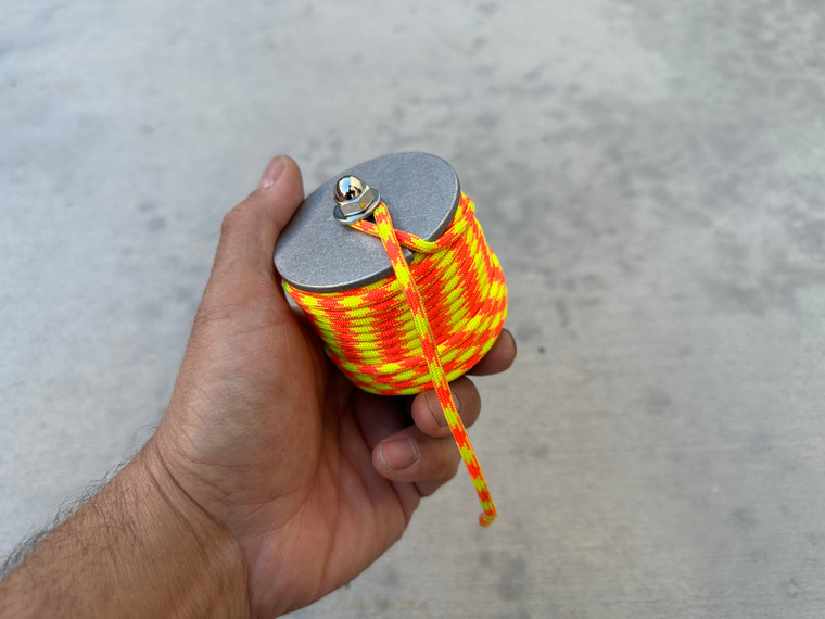 Hand holding a compact bolt-style spool wrapped with orange and yellow paracord.