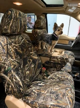 A small dog stands on the center console of a vehicle equipped with custom-fit Realtree Waterproof Camo seat covers, highlighting their pet-friendly durability and rugged style.