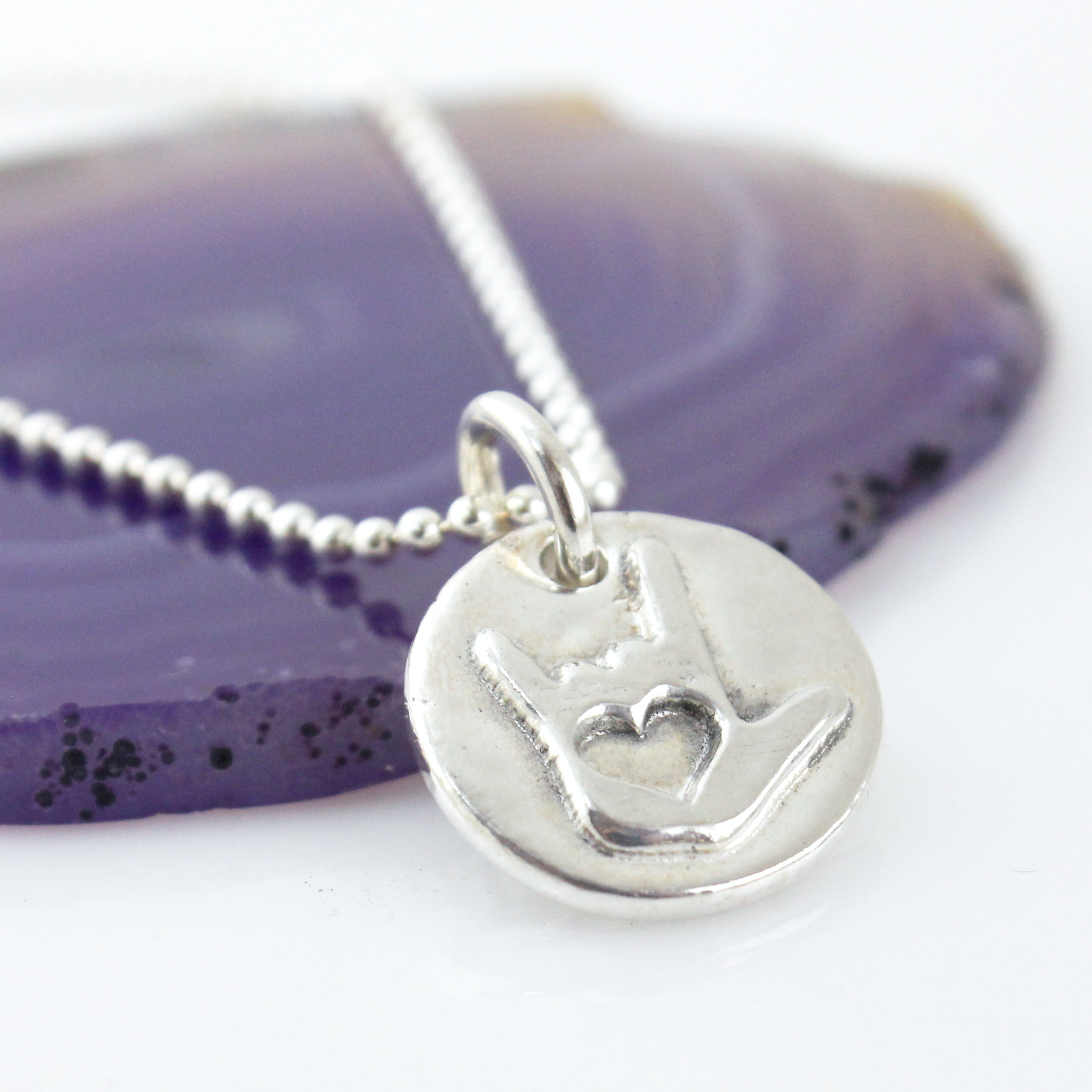 Fine silver ASL I Love You Symbol Token Charm Necklace shown on a sterling ball chain laying partially on a purple colored geode slice.
