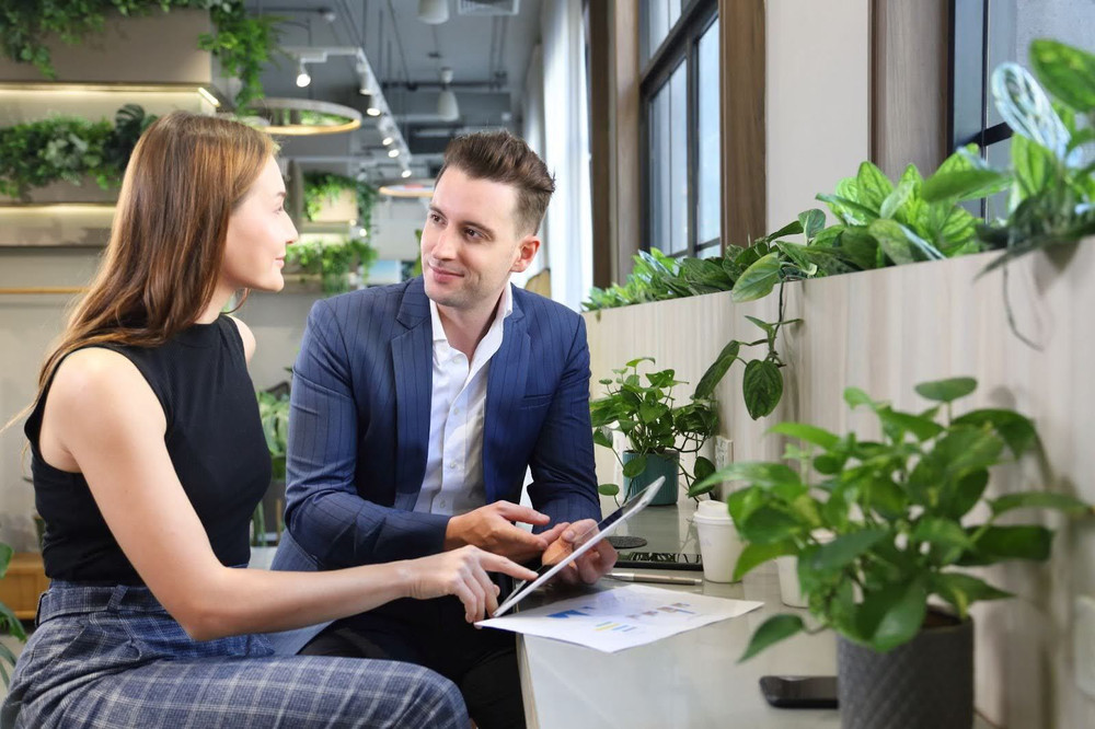 A man and a woman talking and using a tablet in an office surrounded by potted plants