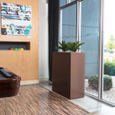 Amesbury tall narrow fiberglass planter box in brown finish with plant against office wall