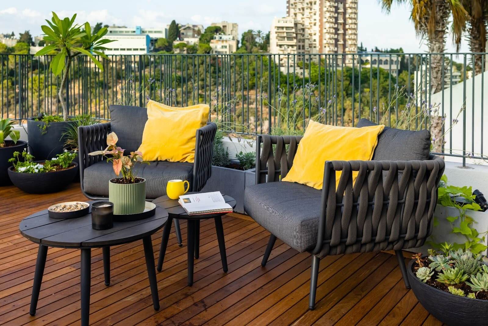 Outdoor patio with two chairs with yellow accents, alongside small tables and potted plants, with a city view in the distance.