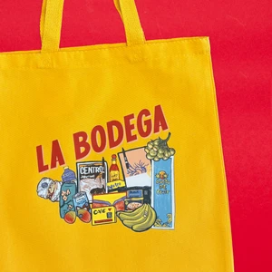 A mustard yellow canvas tote with the words "La Bodega" printed on it showing queso, plátanos, sazón, cafe, mangos, black beans, hot sauce, leche de coco, and more.