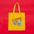 A mustard yellow canvas tote with the words "La Bodega" printed on it showing queso, plátanos, sazón, cafe, mangos, black beans, hot sauce, leche de coco, and more.