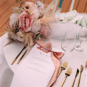 A personalised white napkin with wedding menu printed in elegant black text, styled with a blush coloured ribbon, laser cut guest name, rose gold cutlery and white flowers. A personalised white napkin with wedding menu printed in elegant black text, styled with a blush coloured ribbon, laser cut guest name, rose gold cutlery and white flowers.