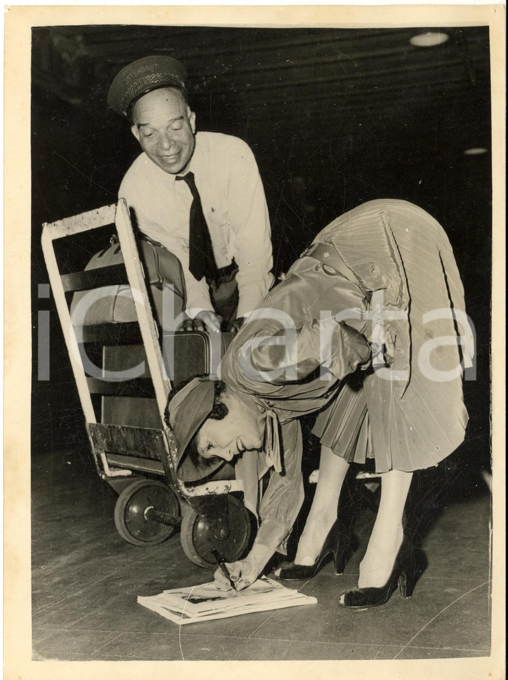 1953 NEW YORK Annette KELLERMAN signing an autograph for a porter *Photo  Fotografia d'epoca con didascalia coeva al verso.   CONDIZIONI: G (ma lievi difetti di stampa) FORMATO: 15x20 cm     originale e autentica 1