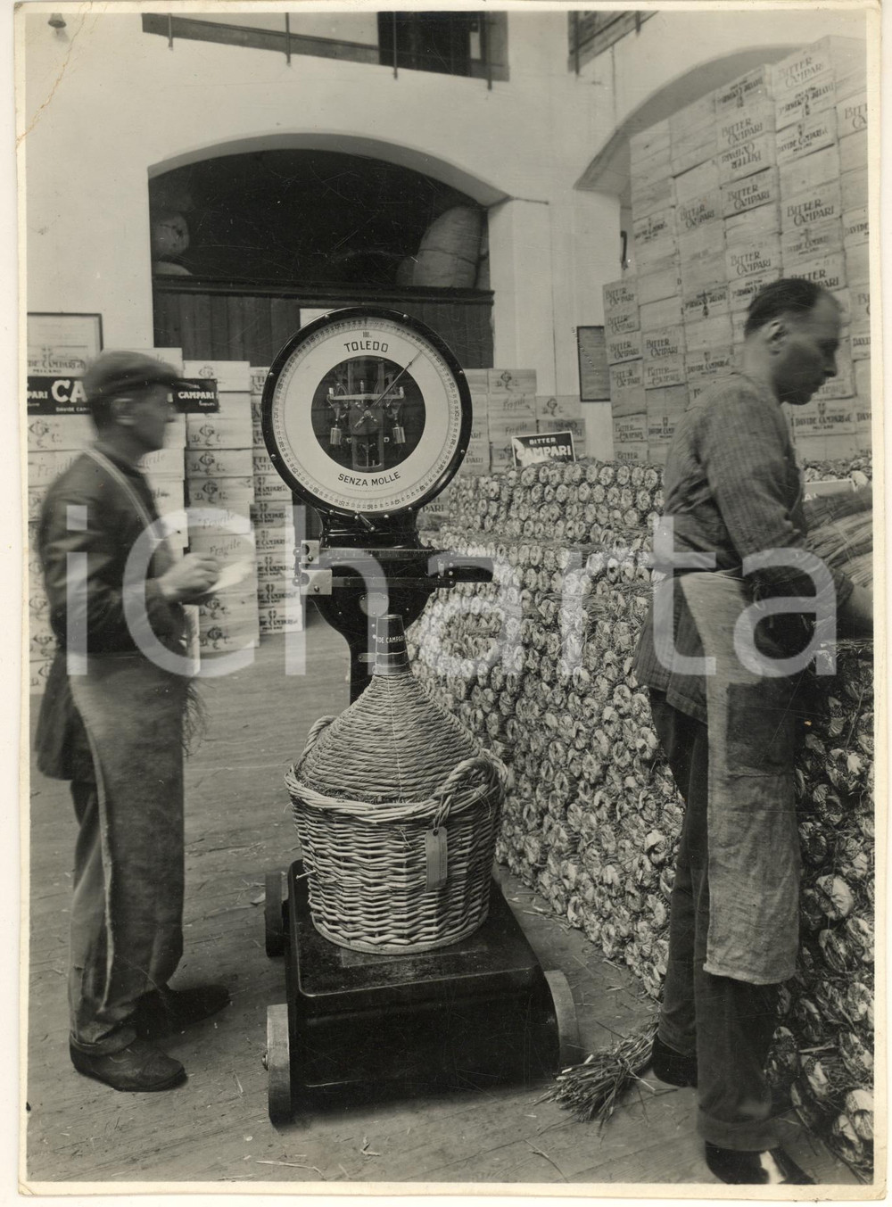 1950 ca ITALIA Magazzino bitter CAMPARI - Peso damigiana con bilancia TOLEDO Fotografia originale d'epoca.  POOR/danneggiato residui di carta e colla al verso; evidenti piegature angolari FORMATO: 17x23 cm originale e autentica 1