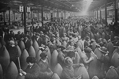 Throngs of women interspersed amongst large artillery shells on a factory floor.