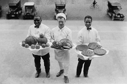 Three Chefs stand on bottom of a line of steps and hold up Thanksgiving platters of Pies, apples and a Turkey