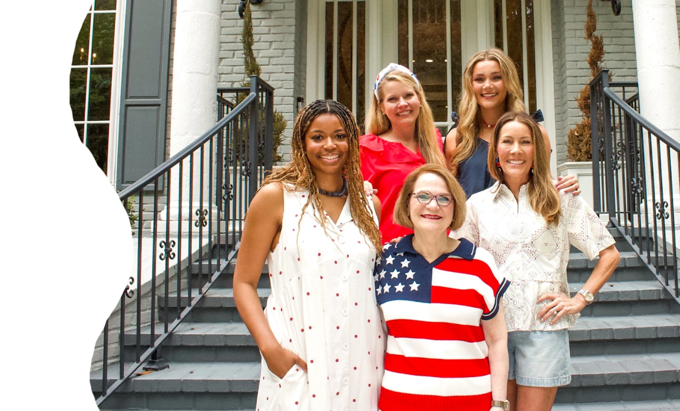 Five women pose together on the front steps of a stately gray brick home with white columns, dressed in red, white, and blue Americana-inspired outfits. The group smiles warmly in front of black iron railings and classic lantern sconces, creating a polish