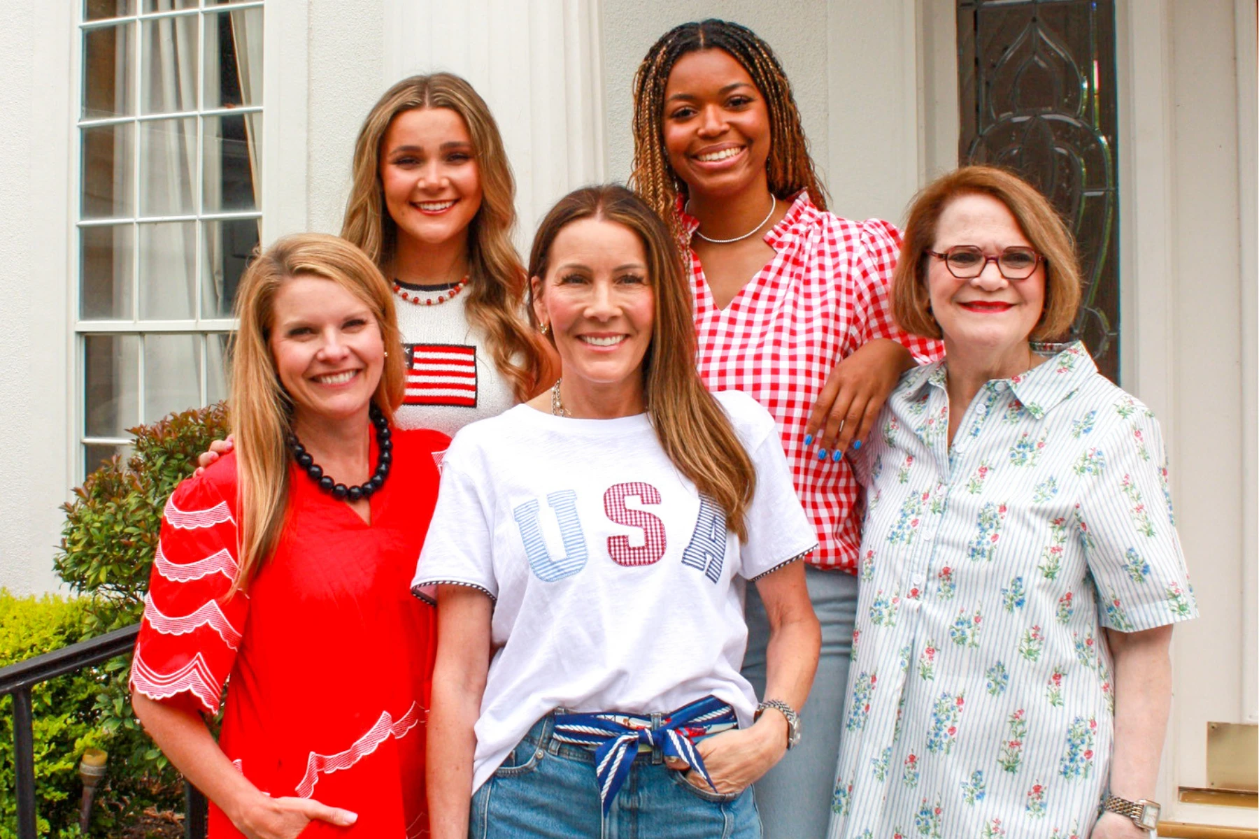 Five women pose together outside a classic white home wearing festive red, white, and blue Americana-inspired outfits. From gingham prints to patriotic graphics and playful summer tops, the group showcases cheerful boutique style perfect for Memorial Day, Fourth of July celebrations, and all-American summer gatherings.