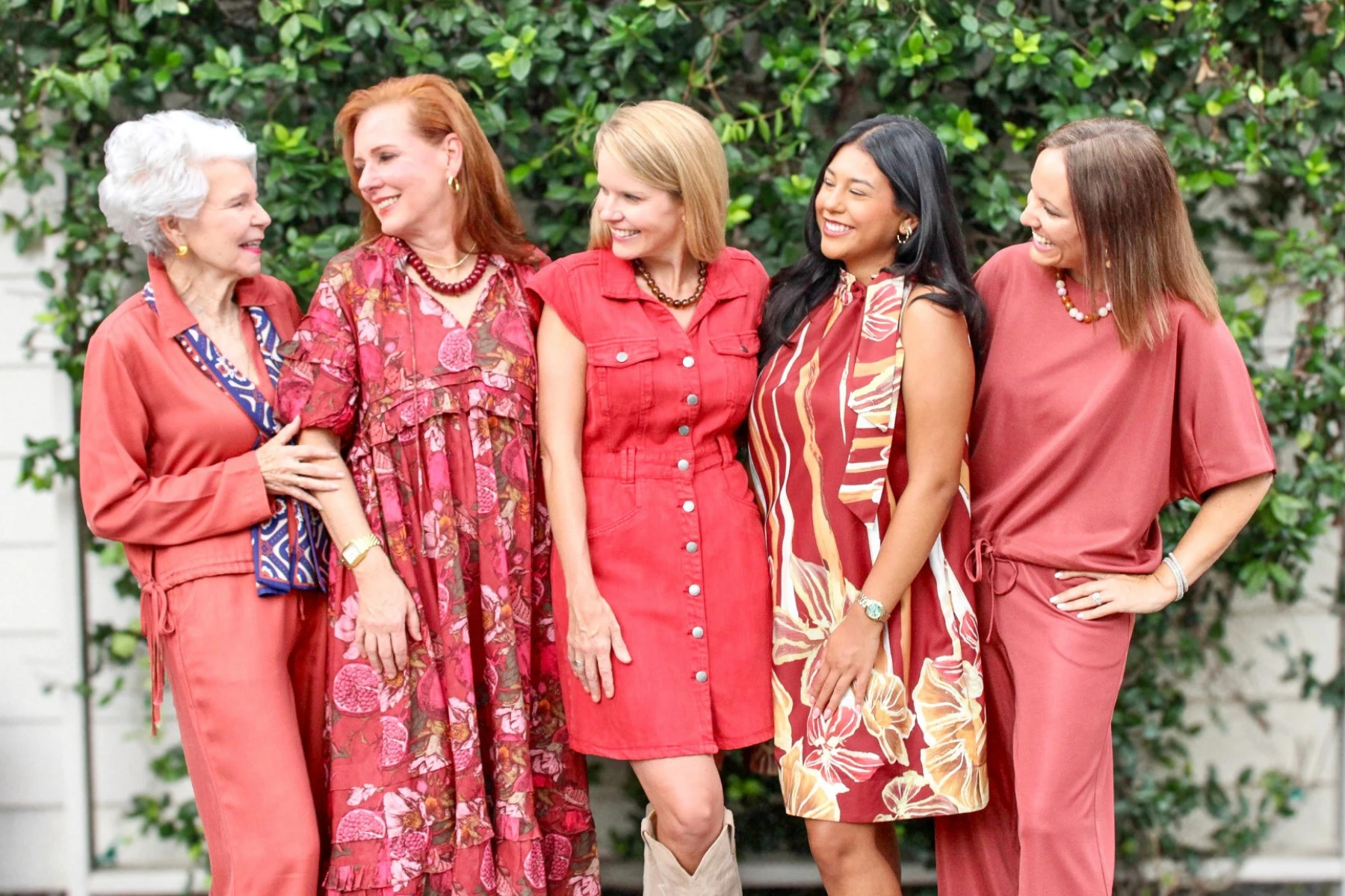 Five women outdoors in coordinating rust and red-toned outfits, mixing floral prints, a denim shirtdress, and solid separates at Magpies Gifts.