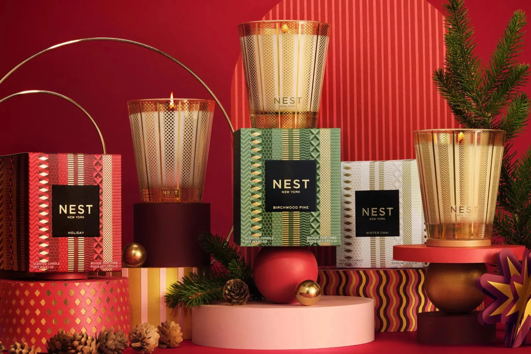 NEST New York holiday candle collection featuring Holiday, Birchwood Pine, and Winter Chai scented candles in elegant gold-patterned glass vessels with festive red, green, and white packaging, surrounded by evergreen branches, pinecones, and gold ornaments against a vibrant red background.