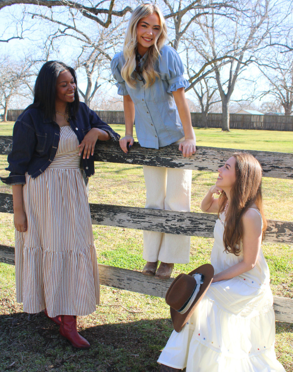 Four women styled in western-inspired outfits gathered around a wooden fence, featuring a striped maxi dress with red boots, a denim blouse with white pants, and a flowing white dress with a brown cowboy hat, set against an open grassy field with bare trees.
