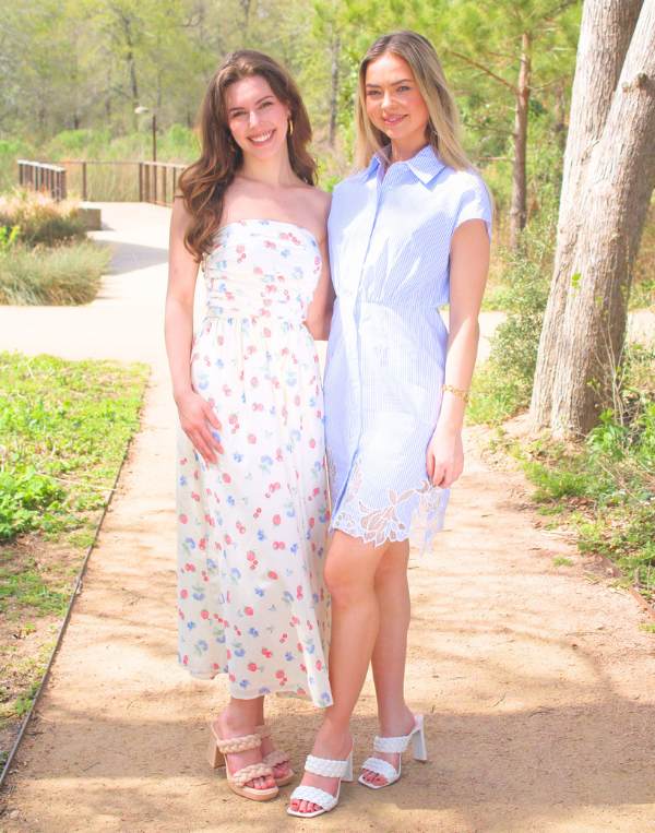 Two women stand side by side on a sunlit outdoor path surrounded by greenery, smiling at the camera. One wears a strapless white maxi dress with a soft floral print and sandals, while the other wears a light blue short-sleeve dress with subtle stripes and lace hem detailing paired with white heels.