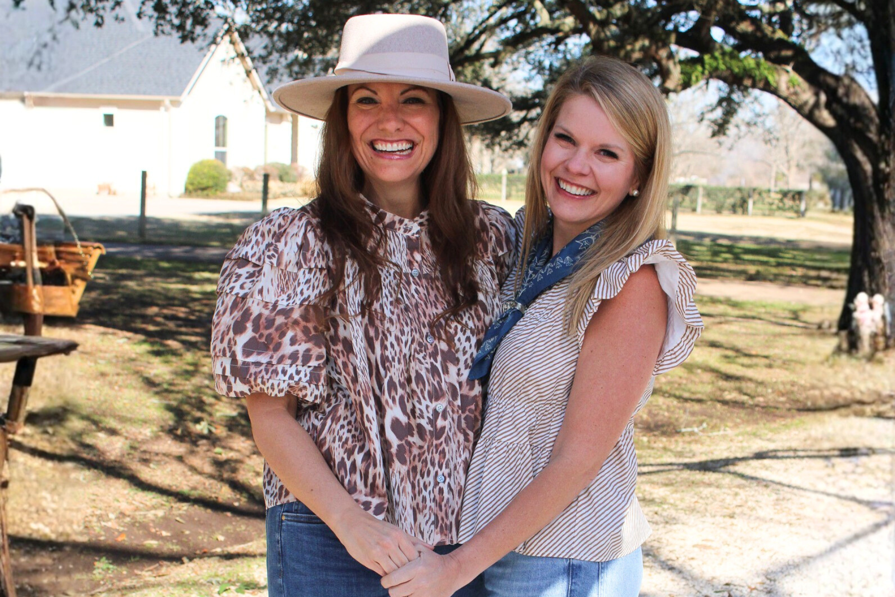 Two women smiling outdoors in rodeo-inspired outfits, one wearing a wide-brim hat and leopard print blouse with jeans, the other in a striped sleeveless top with a blue neck scarf, standing in front of oak trees and a ranch-style home.