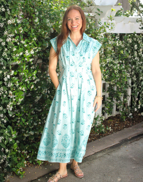 A smiling woman stands outdoors in front of lush green vines with small white flowers, wearing a light blue midi dress with a delicate floral print and cap sleeves. The dress features a button-front bodice, a fitted waist, and a flowy skirt with a decorative border near the hem. She pairs the look with tan strappy sandals and simple jewelry, creating a fresh, effortless spring outfit.