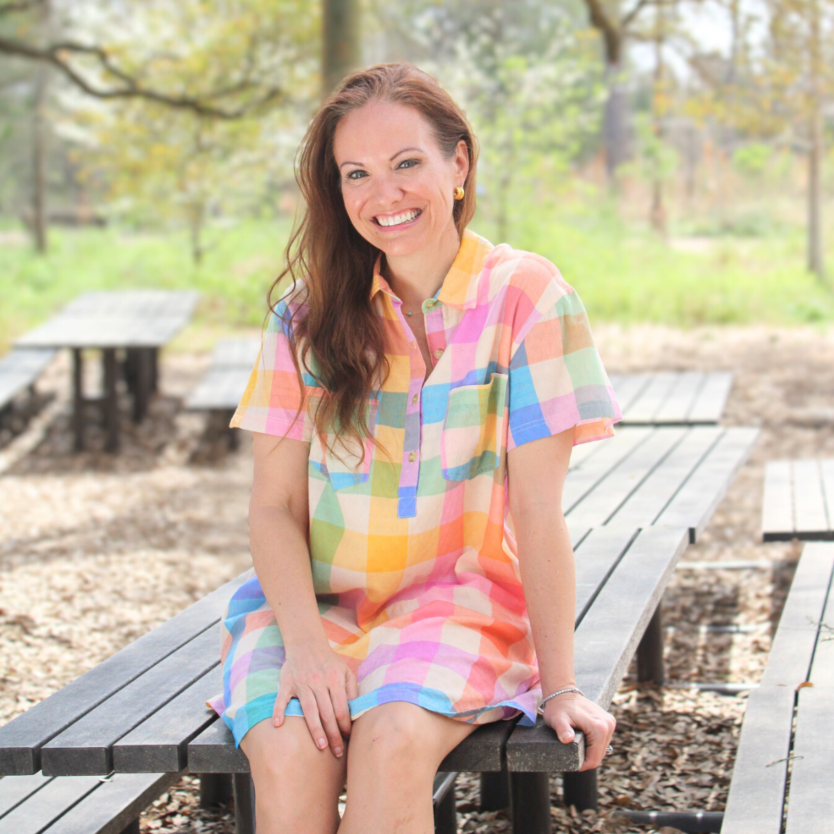 A smiling woman sits on a wooden picnic table outdoors wearing a colorful pastel plaid shirt dress with short sleeves and a button-front neckline. The lightweight dress features soft shades of pink, yellow, blue, and green, creating a cheerful spring look. She pairs the outfit with simple earrings and sandals, surrounded by a natural park setting with trees and soft greenery in the background.