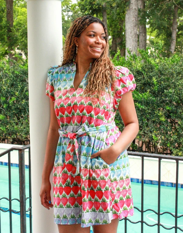 A smiling woman stands by a white column overlooking a pool, wearing a colorful strawberry print romper with flutter sleeves, a tie waist, and pockets. Surrounded by lush greenery, the playful red, pink, and blue fruit pattern creates a fresh and cheerful summer look perfect for warm-weather days and vacation style.