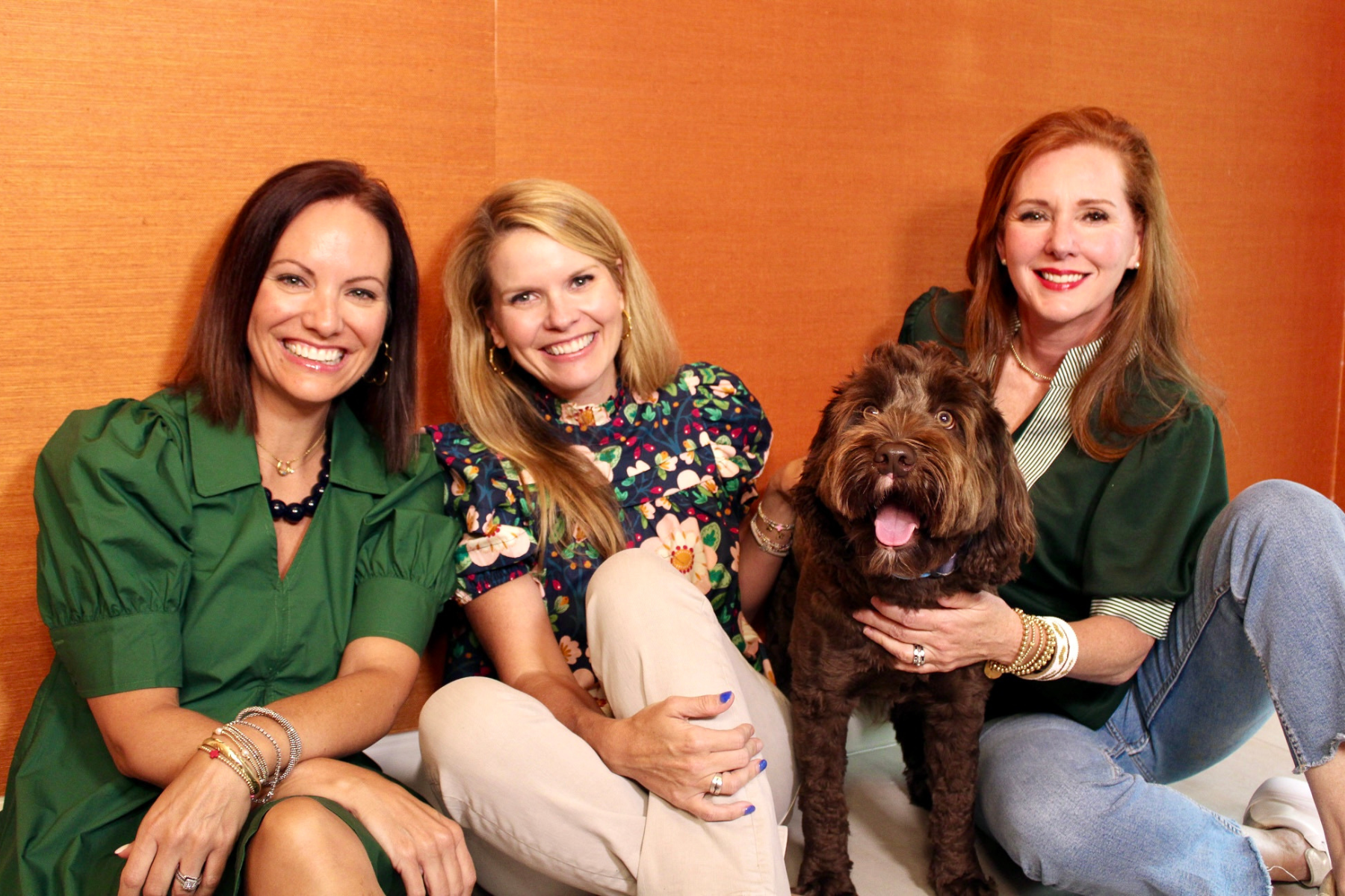 Three women sitting together against an orange background wearing transitional fall outfits — a forest green puff sleeve mini dress, a navy blue floral puff sleeve top with khaki pants, and a green top with a green and white striped shirt with jeans — alongside a fluffy chocolate brown dog.
