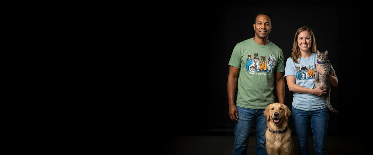 A man in a green dog tee and a woman in a blue cat tee holding a cat with a dog standing below between them on a black background.
