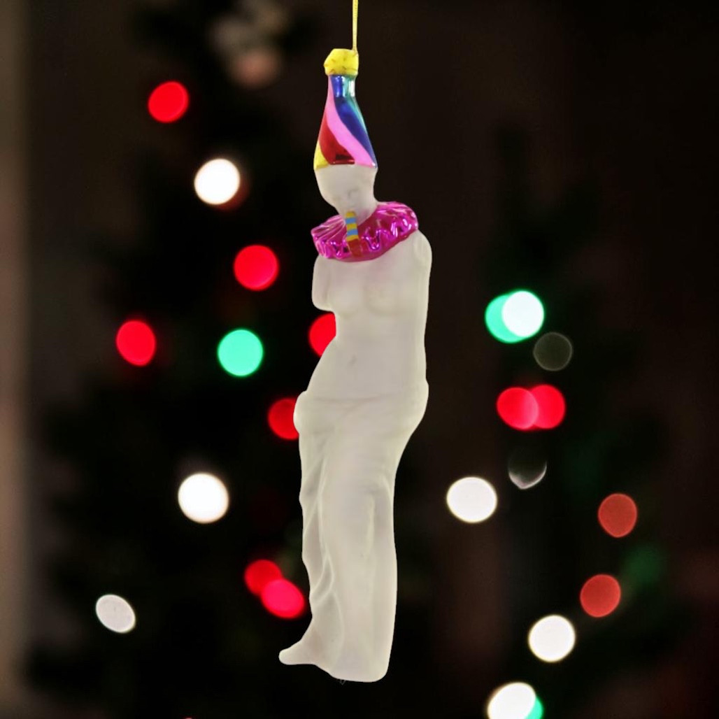 La Fete Venus glass ornament with statue-inspired figure and party hat.
Hanging glass ornament with colorful cone hat and shiny pink collar.
Tall art-inspired glass ornament measuring 1.75 x 1.5 x 7 inches.