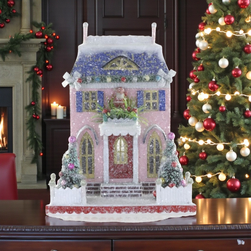Pink glittered cardboard Santa cottage with blue snowy roof, shutters, and white fence.
Christmas village cottage with Santa above the doorway, decorated trees, and frosted sparkle.
Pastel holiday cottage decoration measuring 8.5 x 6.5 x 11 inches.