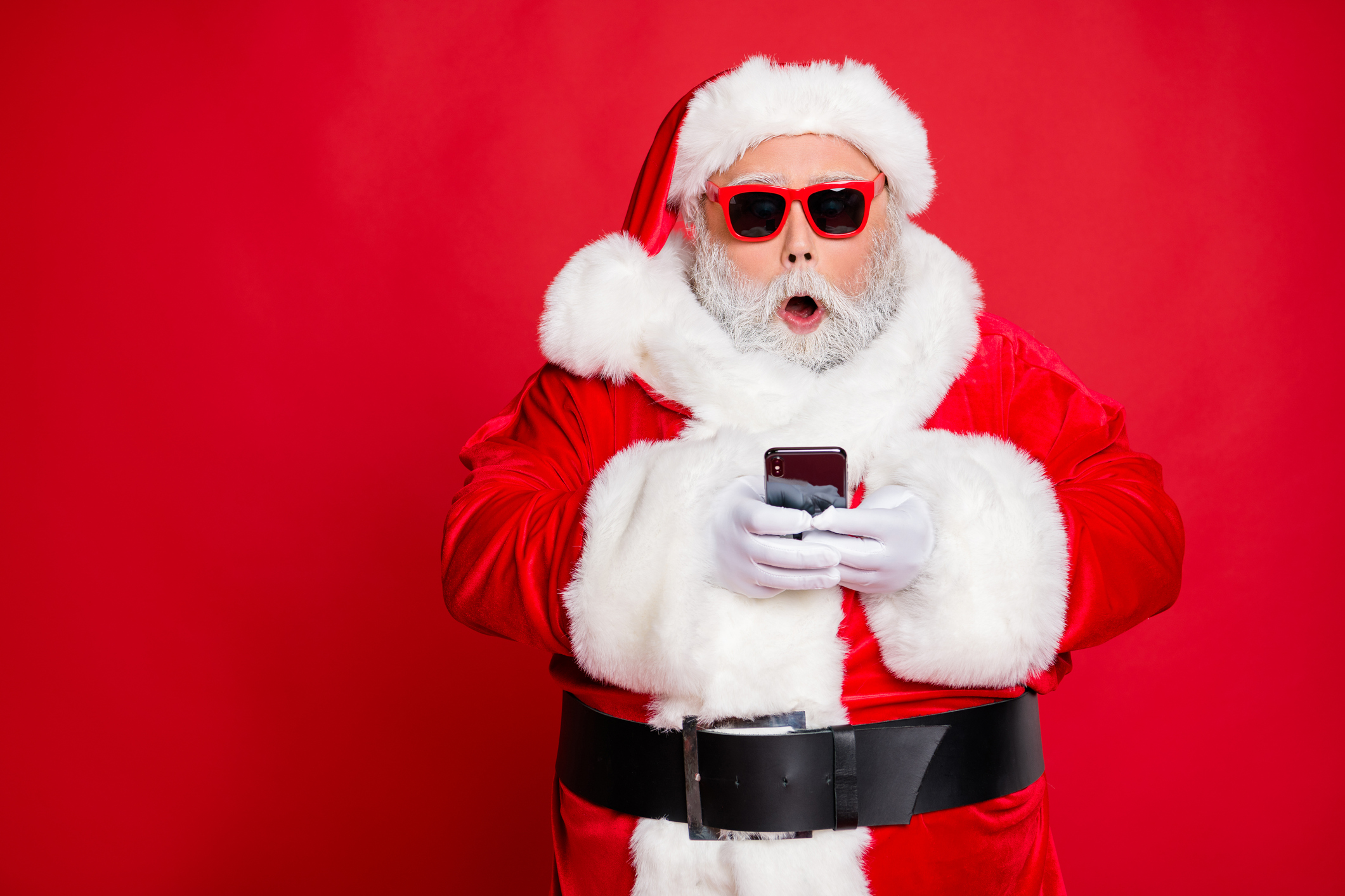 Red background with Santa Clause wearing red thick sunglasses and looking at his smartphone with a surprised look on his face
