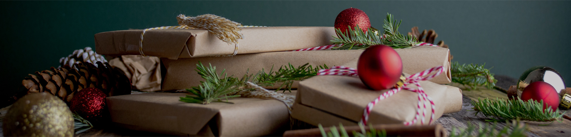 Stylized image of books wrapped in brown paper wrapped wit ribbon piled on a table top surrounded by pine branches, pinecones, and ornaments