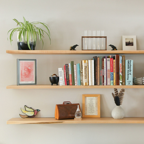 Three long wooden wall-mounted shelves displaying books, framed art, plants, and decorative items in a home. Three long wooden wall-mounted shelves displaying books, framed art, plants, and decorative items in a home.