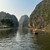 A small wooden rowing boat glides along a calm emerald river winding between towering, jungle-covered limestone karst peaks at Ninh Binh, Vietnam, with more boats in the distance — illustrating a gentle, family-friendly highlight for the July school holidays