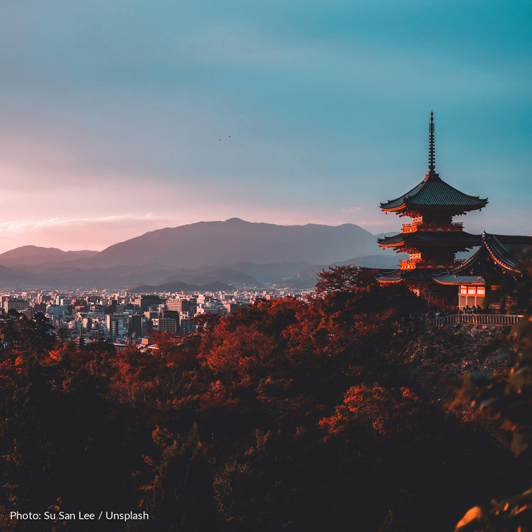 pagoda-in-maples — The tiered pagoda of Kiyomizu-dera standing above Kyoto at dusk, the city and its ring of hills stretching away behind it, with maples in deep autumn red filling the slope in front.