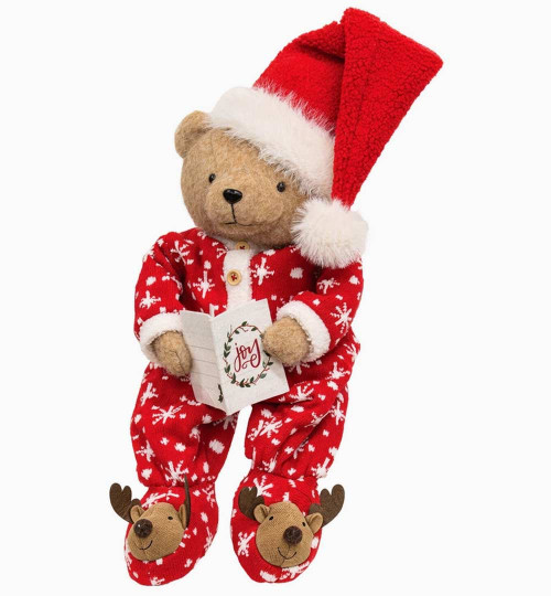 Christmas teddy bear wearing red snowflake pajamas, oversized Santa hat and reindeer slippers, holding a holiday card with the word Joy.
