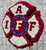 Angled view of a handcrafted firefighter Maltese Cross floral tribute showing sculpted red and white carnation panels and a royal blue floral ring around the IAFF insignia.
