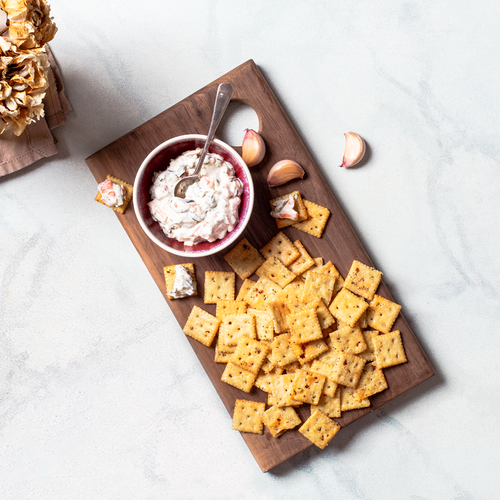 Small cocktail crackers scattered on wooden board with bowl of dip and flowers