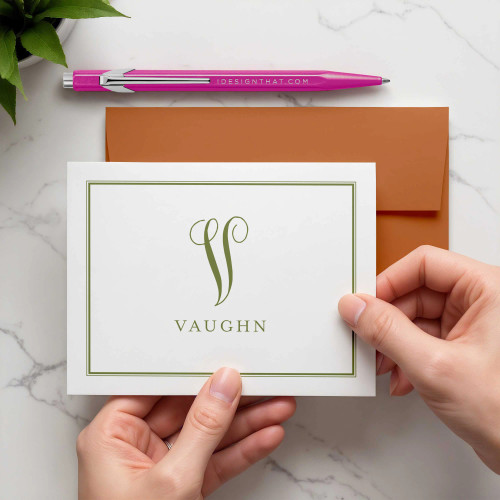 Family Monogram folded notecard showing green initial, surname, and border held above an orange envelope.